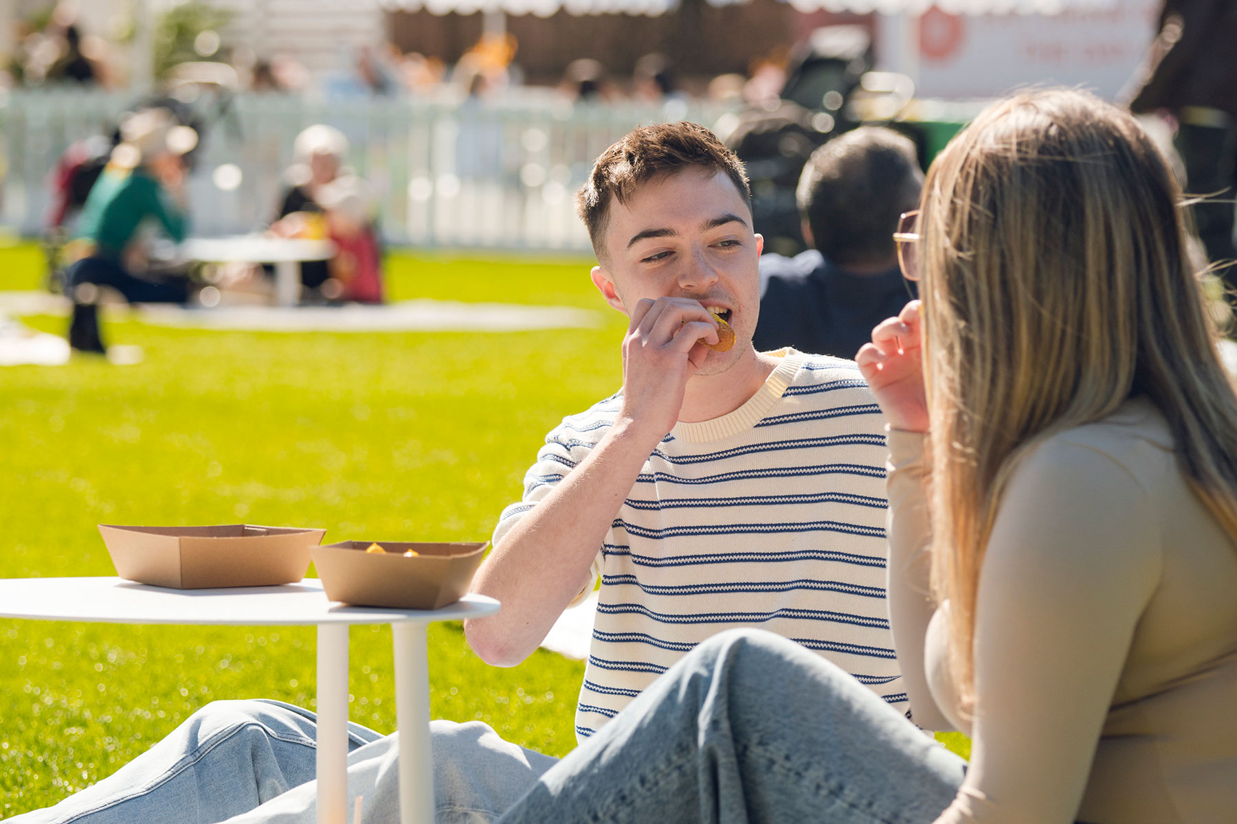 Two people eating sat on a picnic rug at a low table on a sunny day with green grass surrounding