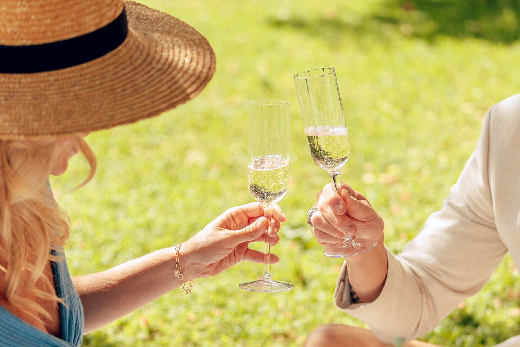 Arms of two people holding glasses of champagne posed to cheers with green grass in the background one person wearing a cane hat