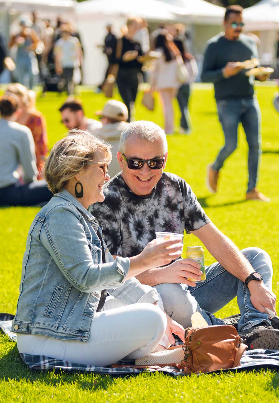 Couple sitting on a picnic rug on green grass smiling enjoying drinks at a festival