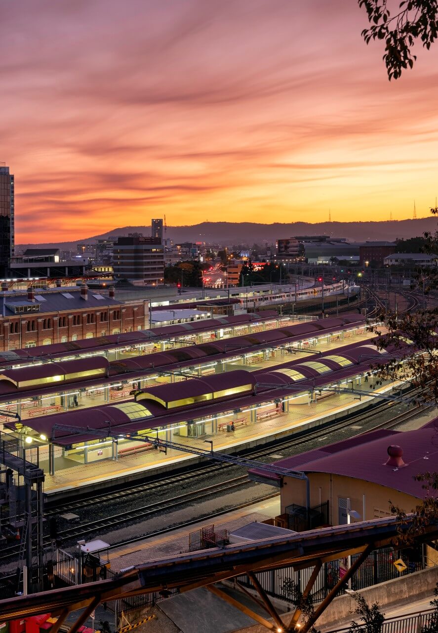 Roma Street Station with illuminated platforms and tracks at sunset, with a glowing orange sky and distant cityscape.